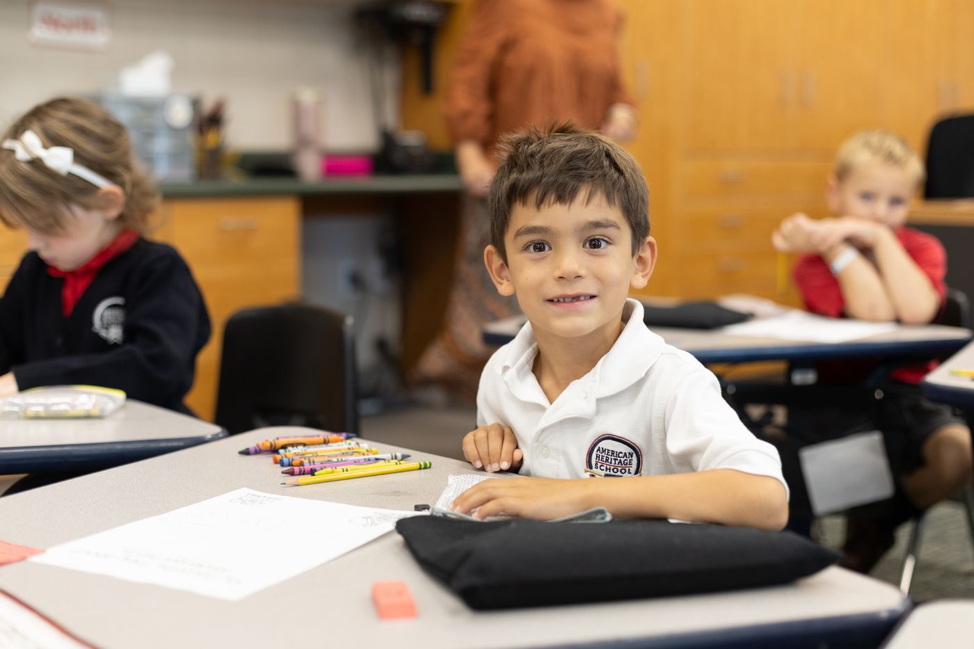 A kindergarten student in an American Heritage School uniform smiles from his desk